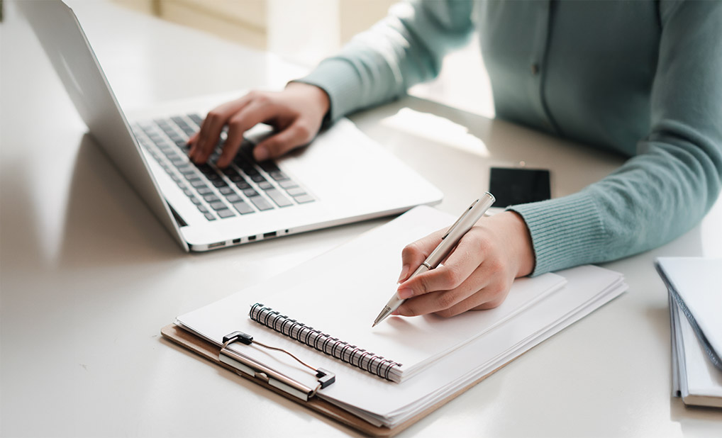 Close up of person typing on laptop and writing in notebook