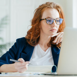 woman looking at a computer and holding a pen above a notepad