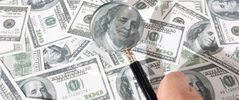 hand holding magnifying glass over pile of $100 bills