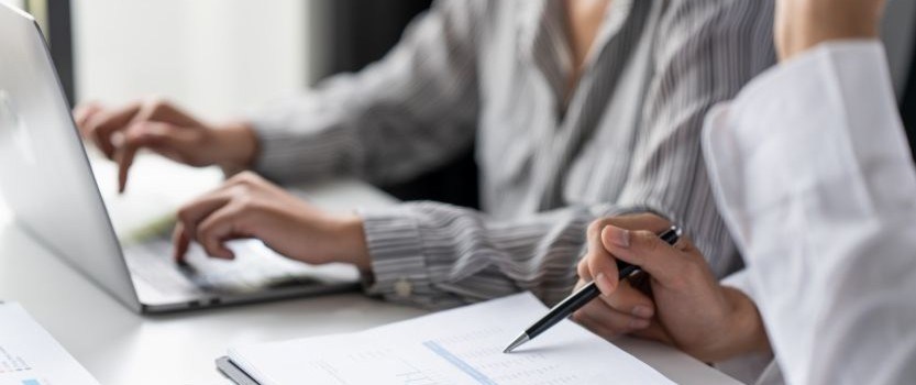 one person writing with pen sitting next to another person typing on a laptop