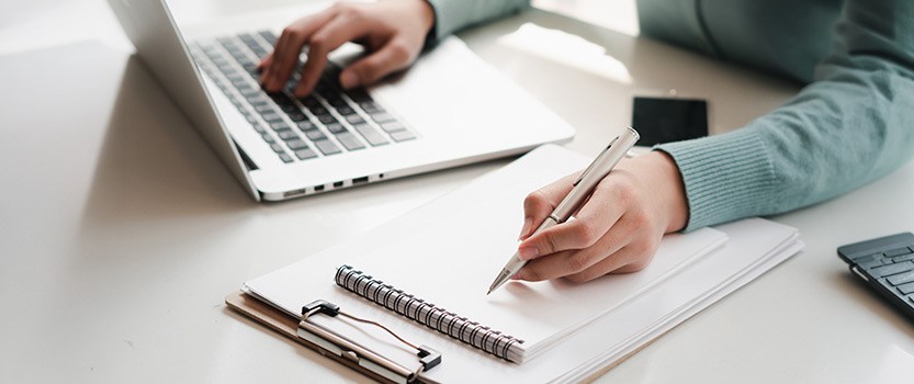 Close up of person typing on laptop and writing in notebook